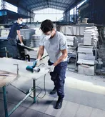 A person wearing safety equipment cuts a stone slab with a power saw in a warehouse.