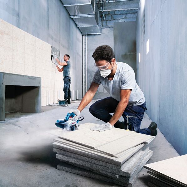 A person wearing safety equipment cuts tiles with a power tool while another installs tiles.
