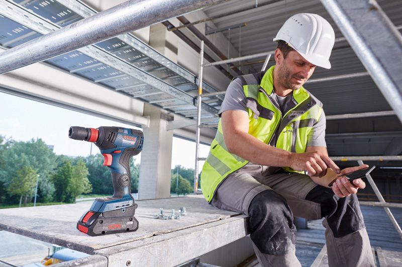 A person wearing safety equipment uses a smartphone beside a cordless drill on a construction site.