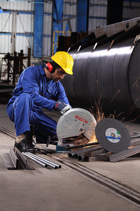 A person wearing safety equipment cuts metal pipes with a chop saw, producing sparks.