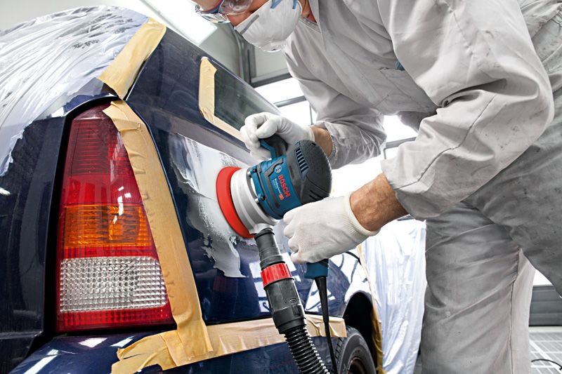 A person wearing safety equipment sands a car’s rear panel with a random orbit sander.