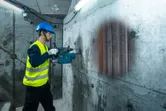 A person wearing safety equipment scans a concrete wall with a detector, revealing pipes inside.