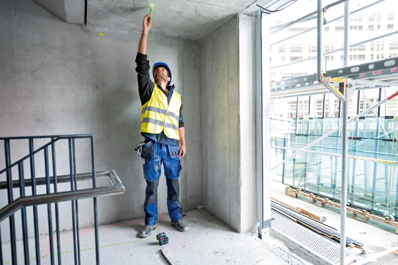A person wearing safety equipment uses a point laser to mark the ceiling in a concrete room.