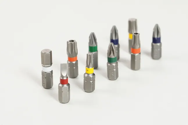 Assorted screwdriver bits with colored bands are arranged on a light surface.