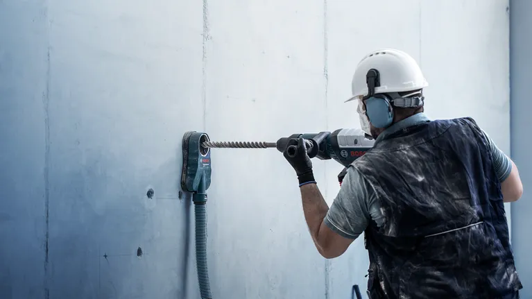 Person wearing safety equipment drills into a concrete wall with a heavy-duty tool.