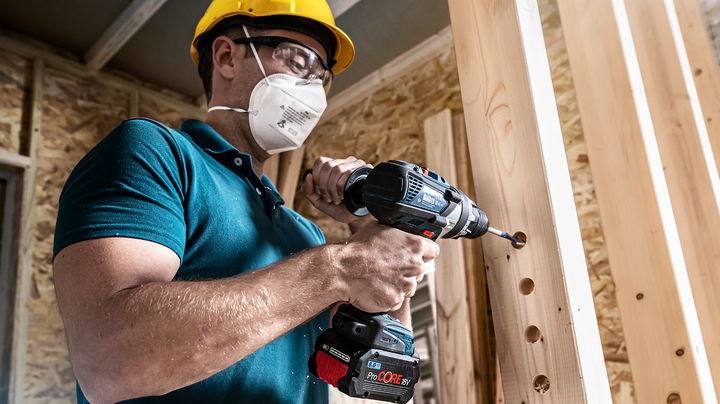 Person wearing safety equipment drills holes in wooden beams.