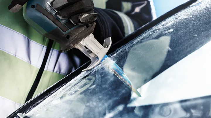 Person wearing safety equipment uses a reciprocating saw to cut a car windshield.