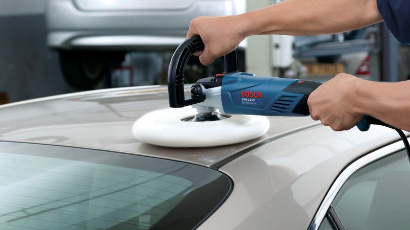 A person polishes a car roof with an electric polisher in a workshop.
