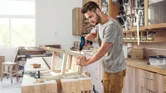 A person drills into a wooden frame on a workbench in a woodworking shop.