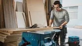 A person wearing safety equipment cuts wood sheets using a table saw in a workshop.