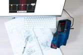 Digital wallscanner detector on architectural blueprints beside a computer keyboard.