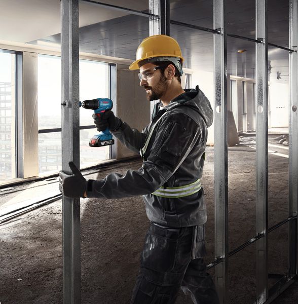A person wearing safety equipment secures metal studs with a cordless drill.