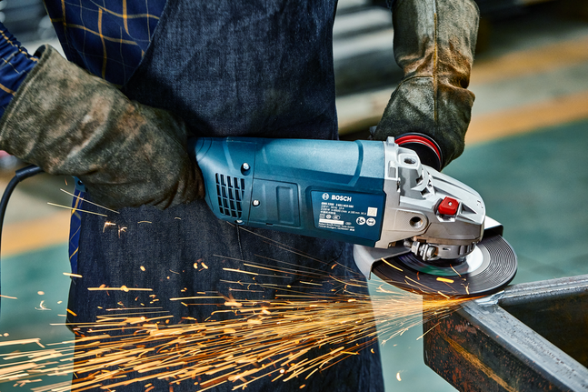 Person wearing safety equipment grinds a metal beam with an angle grinder, producing sparks.