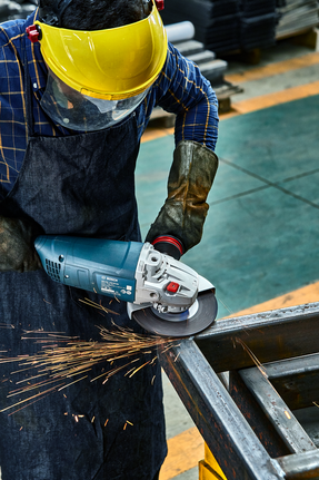 Person wearing safety equipment grinds a metal beam with an angle grinder, sparks flying.