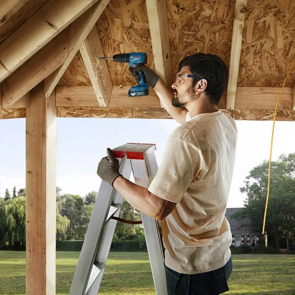 A person wearing safety equipment drills into a wooden beam while standing on a ladder.