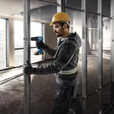 A person wearing safety equipment uses a cordless drill on a metal wall frame.