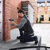 Person wearing safety equipment drills into a brick wall outdoors.