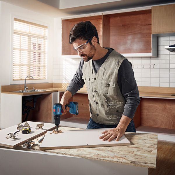 A person wearing safety equipment drills into a wooden panel in a kitchen workspace.