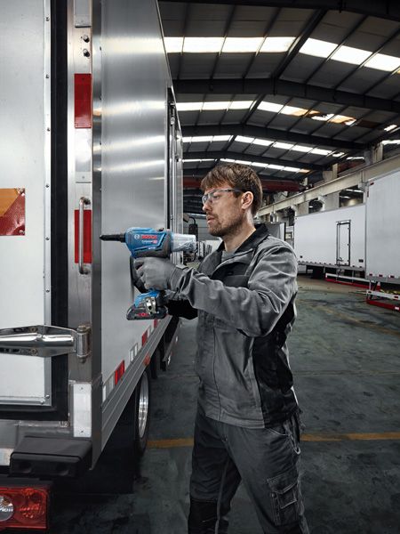 A worker wearing safety equipment uses a cordless drill to fasten metal on a trailer.