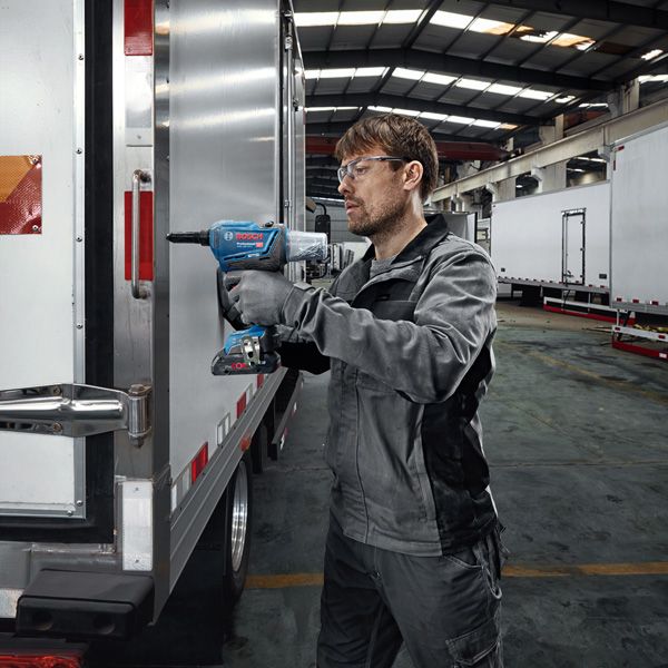 A person wearing safety equipment uses a cordless impact wrench on a truck door.