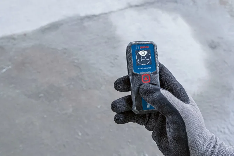 Person wearing safety equipment operates a compact laser measure on a concrete surface.