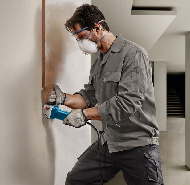 Person wearing safety equipment uses an angle grinder to cut a vertical groove in a wall.