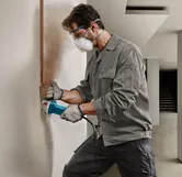 Person wearing safety equipment uses an angle grinder to cut a vertical groove in a wall.
