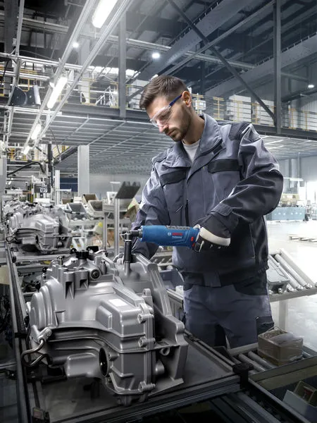 A person wearing safety equipment uses a power tool to assemble machinery in a factory.