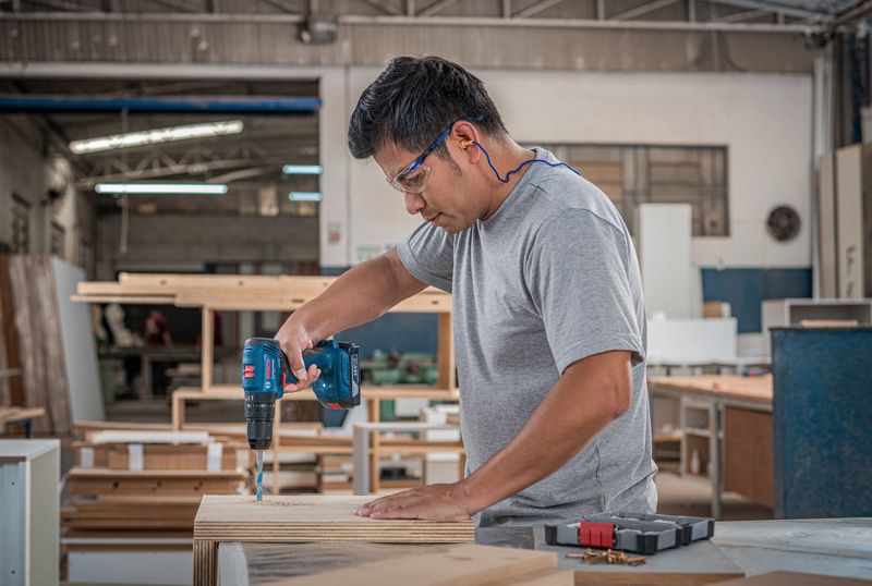 A person wearing safety equipment drills into wood on a workbench.
