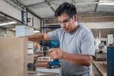 A person wearing safety equipment drills into plywood in a woodworking shop.