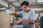 A person wearing safety equipment drills into plywood in a woodworking shop.