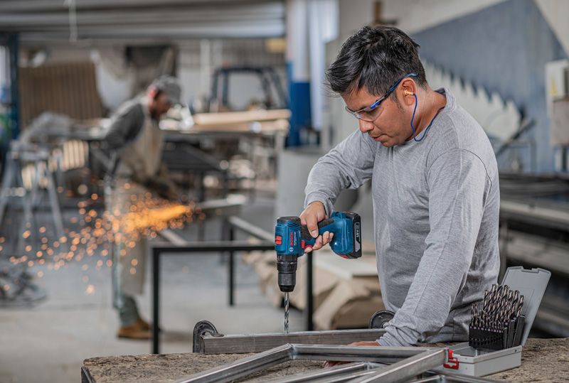 A person wearing safety equipment drills metal, with another worker grinding in the background.