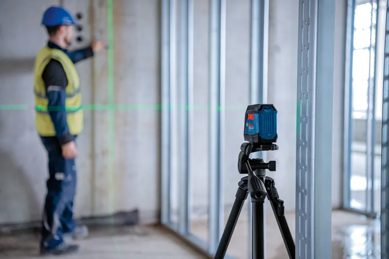 Person wearing safety equipment aligns framing studs using a laser leveling tool on a tripod.