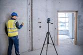 A person wearing safety equipment marks a concrete wall using a laser leveling tool on a tripod.