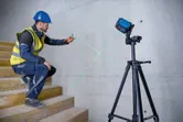 A person wearing safety equipment marks a wall using a green laser leveling tool on a tripod.