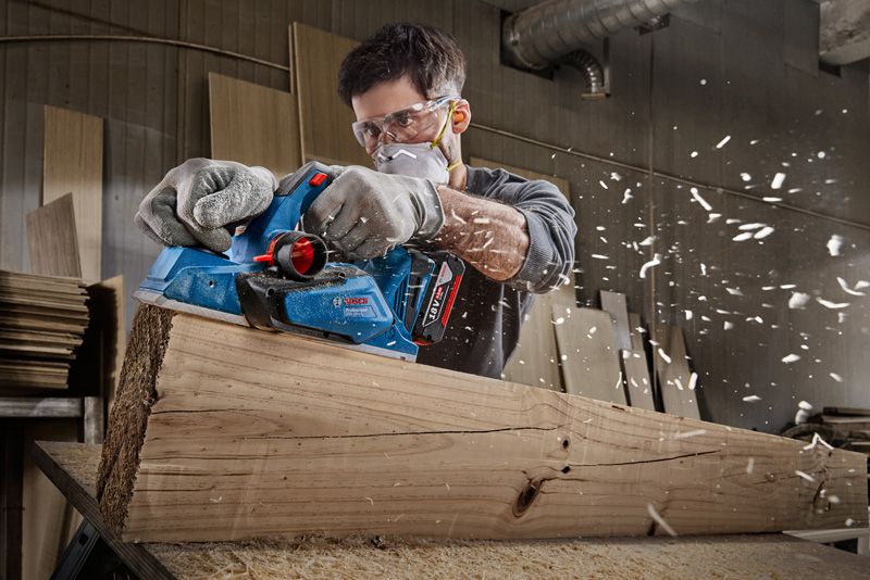 A person wearing safety equipment planes a wooden beam with an electric planer.