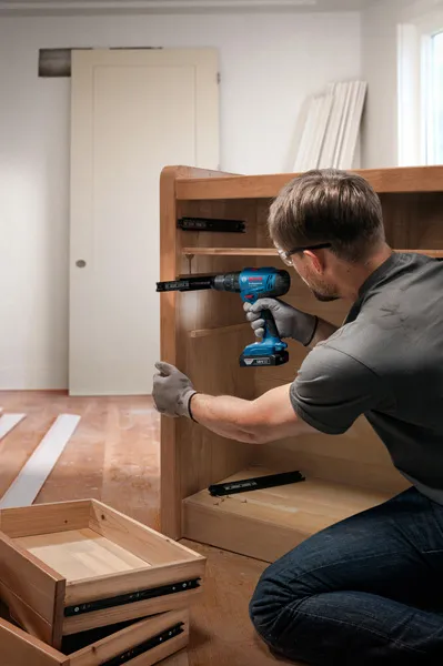 Person wearing safety equipment installs cabinet drawer slides using a cordless drill.