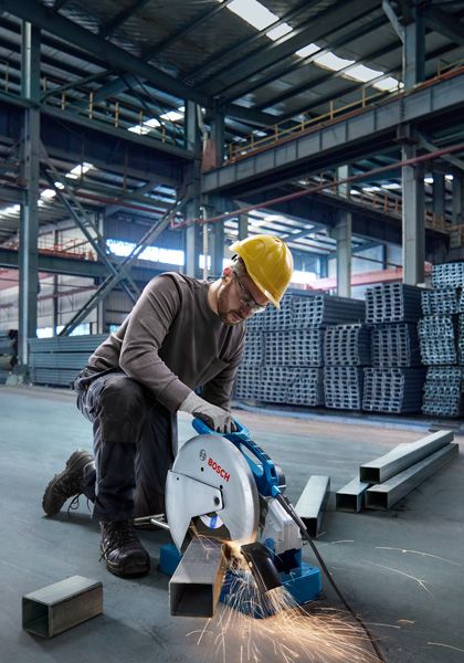 A person wearing safety equipment cuts metal pipes with a power saw in a warehouse.