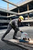 A person wearing safety equipment operates a chop saw, cutting metal at a construction site.