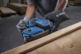 Person wearing safety equipment sanding wood with a blue belt sander in a workshop.