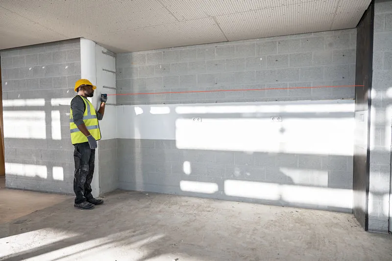 A person wearing safety equipment uses a laser leveling tool to project a straight line on a wall.