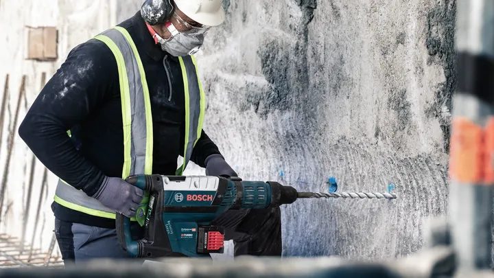 A person wearing safety equipment drills into concrete with a rotary hammer.