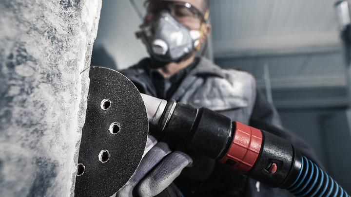 A person wearing safety equipment sands a concrete wall with a power tool.