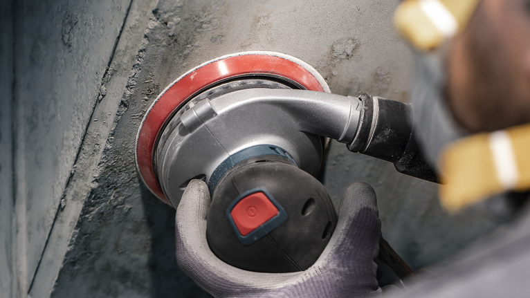 Person wearing safety equipment uses a power sander on a concrete wall.