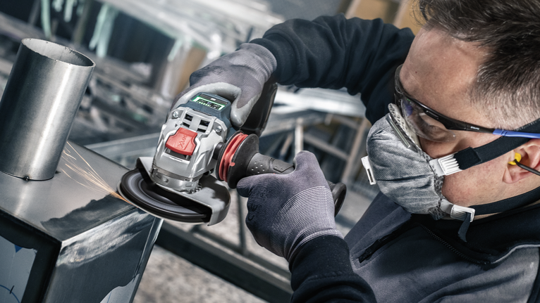 A person wearing safety equipment grinds metal with a power tool.