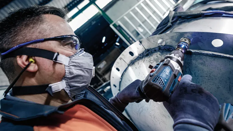 A person wearing safety equipment drills into a large metal cylinder.