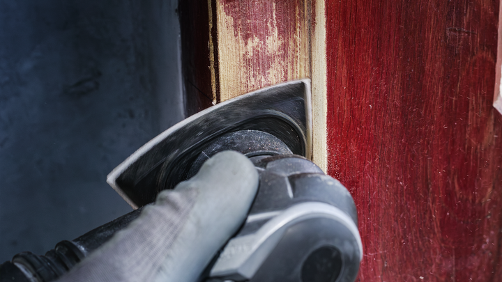 Person wearing safety equipment sands paint off a wooden door with a power tool.