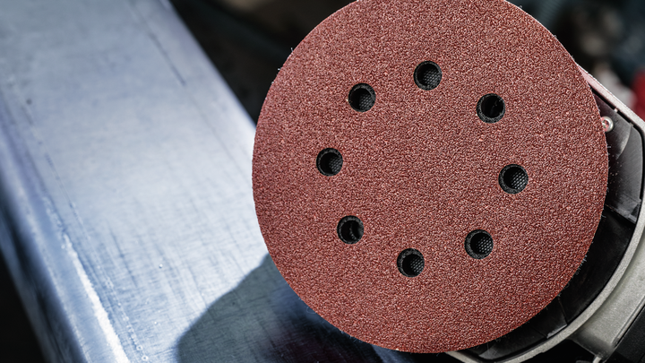 A disc sander with a red abrasive pad is positioned above a metal surface.