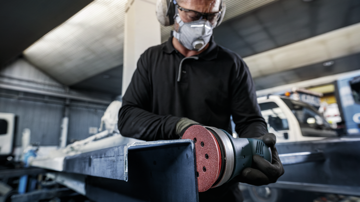 Person wearing safety equipment sands a metal beam in an industrial workshop.