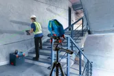 A person wearing safety equipment aligns a tool on a staircase using a laser leveling tool.
